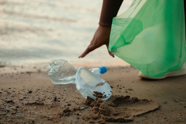 Mulher africana irreconhecível coletando lixo derramado de areia na praia em saco de plástico verde Poluição ecológica