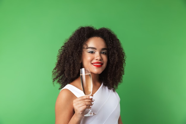 Mulher africana jovem alegre usando vestido comemorando isolado, bebendo champanhe em uma taça