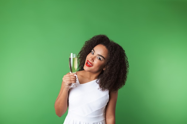 Mulher africana jovem alegre usando vestido comemorando isolado, bebendo champanhe em uma taça