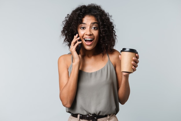 Mulher africana jovem e feliz, vestida de maneira casual, isolada, falando no celular, segurando uma xícara de café para viagem