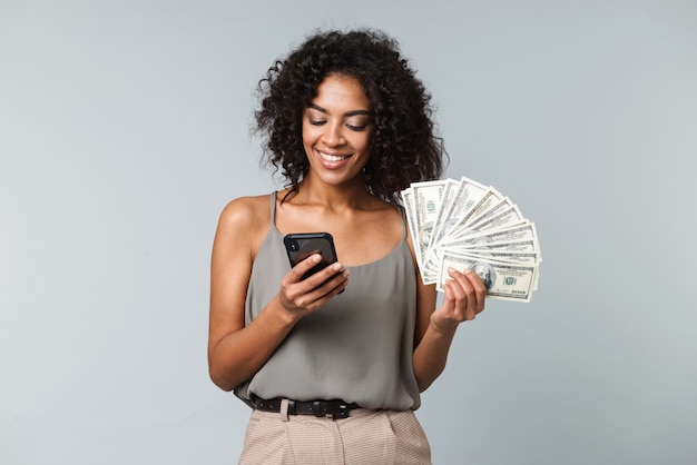 Mulher africana jovem feliz isolada, segurando um monte de notas de dinheiro, usando telefone celular