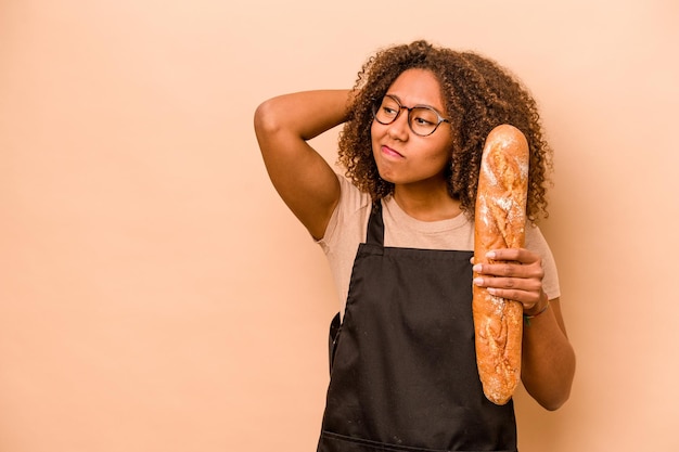 Mulher africana jovem padeiro segurando um pedaço de pão isolado em fundo bege tocando a parte de trás da cabeça pensando e fazendo uma escolha