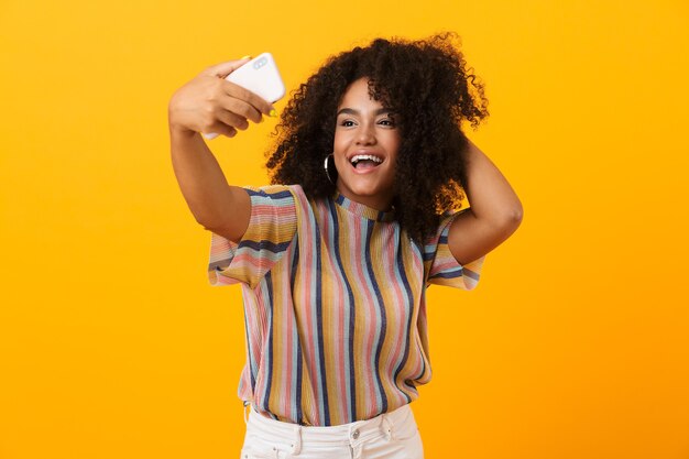 Mulher africana posando isolada sobre o espaço amarelo, tome uma selfie pelo celular.