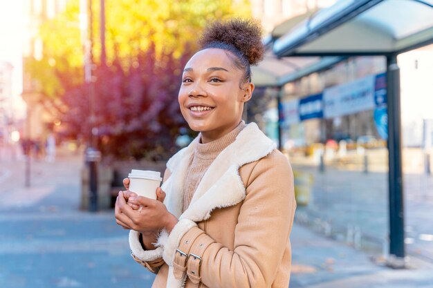 Mulher africana segura xícara de café Uma sorridente morena encaracolada de suéter marrom esperando um bonde