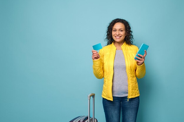 Mulher africana sorridente em uma jaqueta amarela fica contra o fundo da parede azul, na frente de sua mala, segura o celular e o cartão de plástico com um espaço de anúncio de cópia vazio. Viagem, reserva, conceito bancário