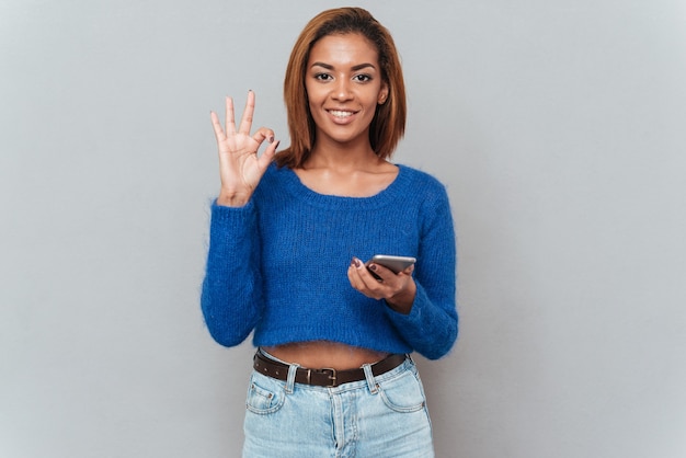 Mulher africana sorridente feliz em suéter e jeans com telefone mostrando sinal de ok. Fundo cinza isolado