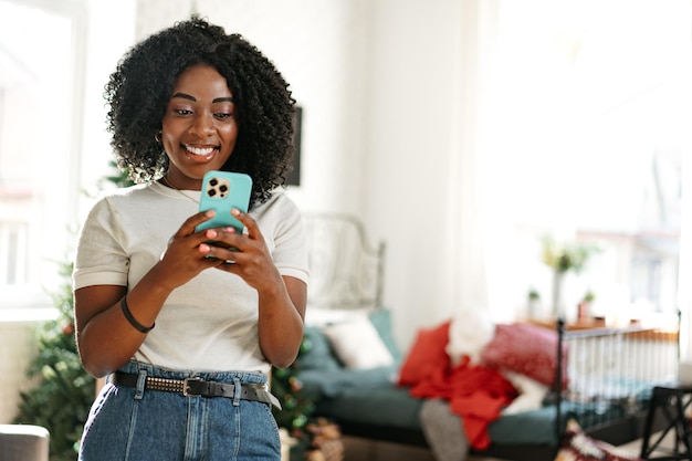 Mulher africana sorridente sentada no sofá em casa e usando smartphone