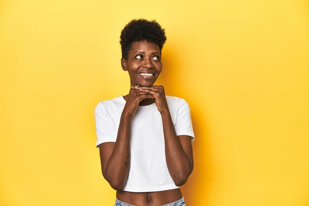 Foto mulher africana t-shirt branco cabelo curto estúdio amarelo mantém as mãos sob o queixo está olhando felizmente de lado