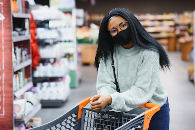 Mulher africana usando máscara médica descartável, compras no supermercado durante o surto de pandemia de coronavírus. época de epidemia.