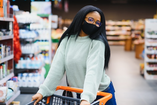 Mulher africana usando máscara médica descartável, compras no supermercado durante o surto de pandemia de coronavírus. Época de epidemia.