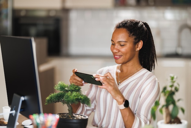 Mulher africana usando seu smartphone para tirar fotos de suas plantas domésticas enquanto faz um vlog sobre cuidados com as plantas em seu escritório em casa.