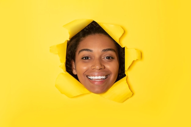 Mulher afro-americana animada posando em fundo de papel amarelo rasgado olhando para a câmera e sorrindo pelo buraco