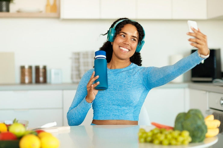 Foto mulher afro-americana atirando selfie e segurando garrafa vestindo roupas esportivas sentada na cozinha após treino doméstico