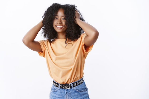 Foto mulher afro-americana atraente e despreocupada vestindo uma camiseta laranja da moda dançando alegremente, sorrindo, tocando o corte de cabelo afro encaracolado encantada, sentindo o cabelo fofo, parede branca em pé