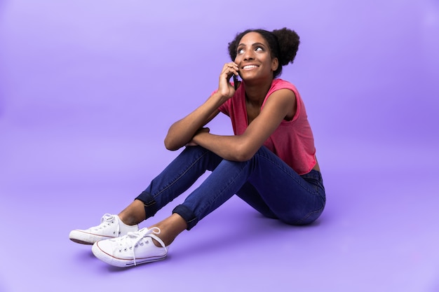 Mulher afro-americana atraente engraçada sorrindo e falando no smartphone, enquanto está sentado no chão, isolado