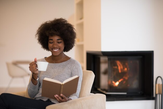 mulher afro-americana bebendo café lendo livro na lareira. Jovem negra com bebida quente relaxante aquecimento aquecimento. outono em casa.