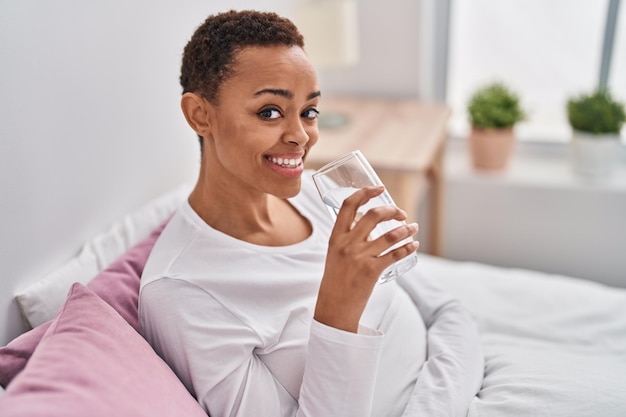 Mulher afro-americana bebendo um copo de água sentado na cama no quarto