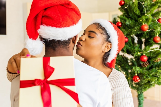 Mulher afro-americana beijando na bochecha do namorado para caixa de presente em casa perto da árvore de natal