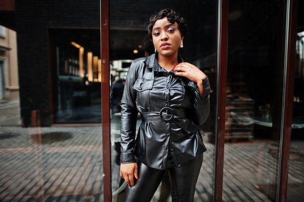 Mulher afro-americana bonita elegante posando de jaqueta de couro preta e calças na rua. Brinco no nariz.