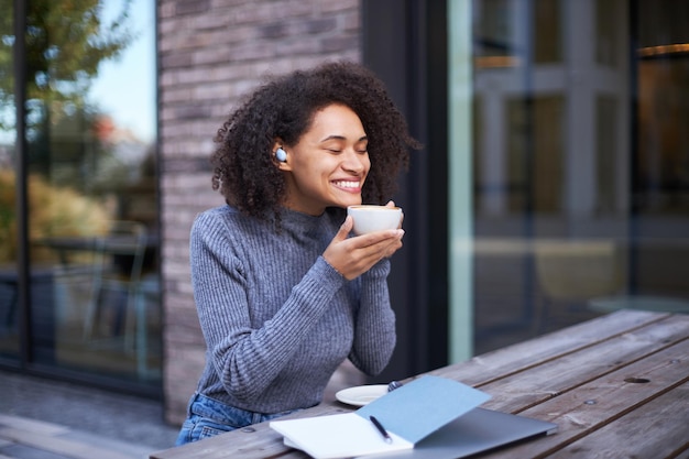 Mulher afro-americana bonita em roupas casuais tomando um gole de café com aroma de hor em um café ao ar livre