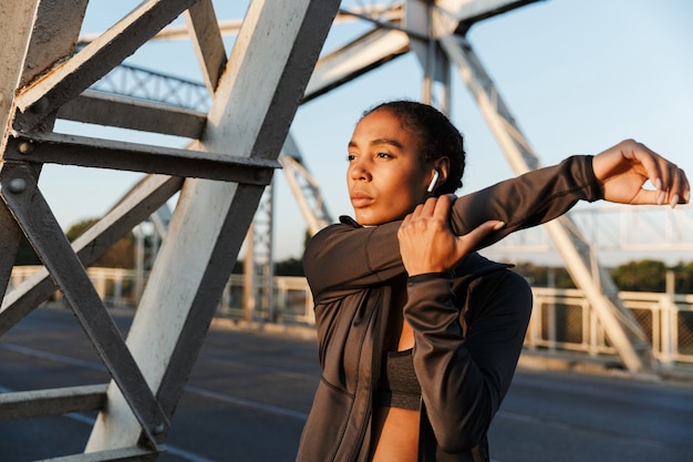 Mulher afro-americana bonita em roupas esportivas, usando fones de ouvido e esticando os braços enquanto se exercita na ponte velha