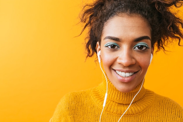 Mulher afro-americana cândida em roupas casuais, ouvindo música com fones de ouvido brancos, isolada