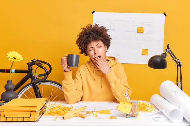 Foto mulher afro-americana cansada e sonolenta se prepara para os exames, bebe café e boceja poses no espaço de coworking. trabalhador de escritório com excesso de trabalho ocupado termina de trabalhar nos esboços do arquiteto. conceito de prazo