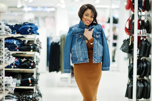 Mulher afro-americana chique em vestido de túnica marrom e jaqueta jeans posou na loja de roupas É hora de fazer compras