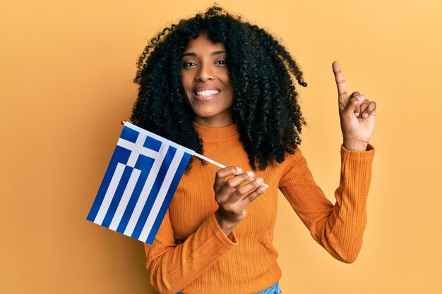 Mulher afro-americana com cabelo afro segurando a bandeira da grécia sorrindo com uma ideia ou pergunta apontando o dedo com cara feliz número um