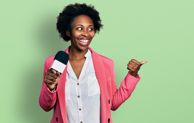Mulher afro-americana com cabelo afro segurando microfone repórter apontando o polegar para o lado sorrindo feliz com a boca aberta