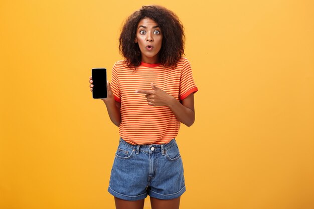 Mulher afro-americana com cabelo encaracolado apontando para o celular com uma expressão de espanto sobre uma parede laranja