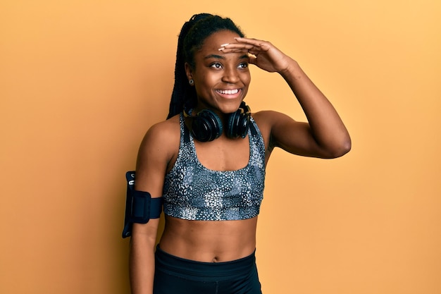 Mulher afro-americana com cabelo trançado vestindo roupas esportivas e faixa de braço muito feliz e sorrindo olhando para longe com a mão sobre a cabeça. conceito de pesquisa.