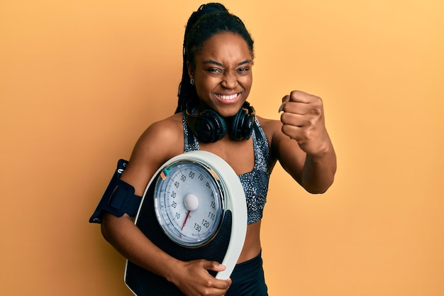 Mulher afro-americana com cabelo trançado vestindo roupas esportivas segurando balança irritada e frustrada gritando de raiva, gritando loucamente de raiva e mão levantada