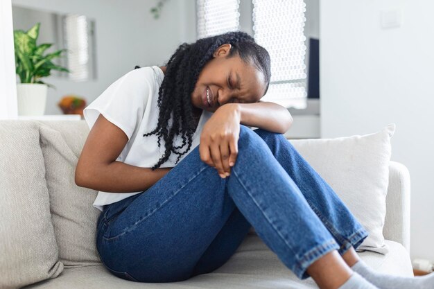 Mulher afro-americana com expressão dolorosa segurando as mãos contra a barriga sofrendo dor do período menstrual deitada triste na cama de casa com cãibra na barriga no conceito de saúde feminina