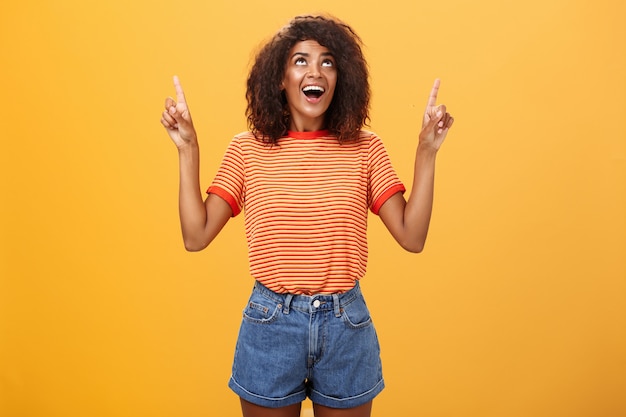 Mulher afro-americana com penteado afro em camiseta e shorts jeans apontando e olhando para cima com olhar interessado sobre a parede laranja