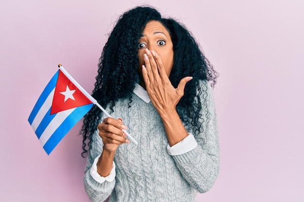 Mulher afro-americana de meia-idade segurando a bandeira de cuba cobrindo a boca com a mão chocada e com medo de erro expressão surpresa