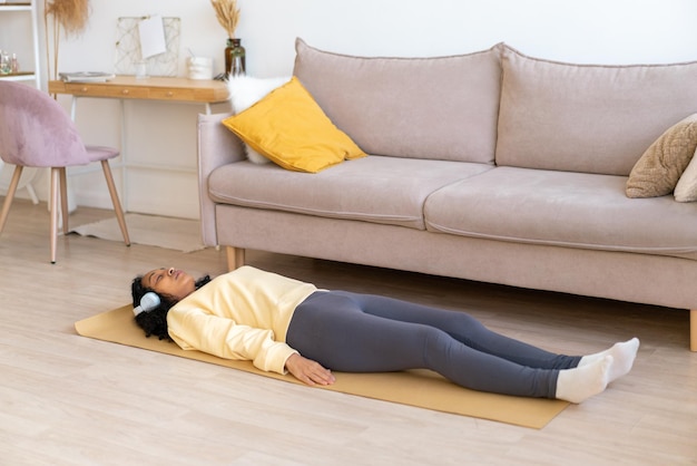 Mulher afro-americana deitada no tapete na sala de estar relaxando depois de fazer exercícios em fones de ouvido