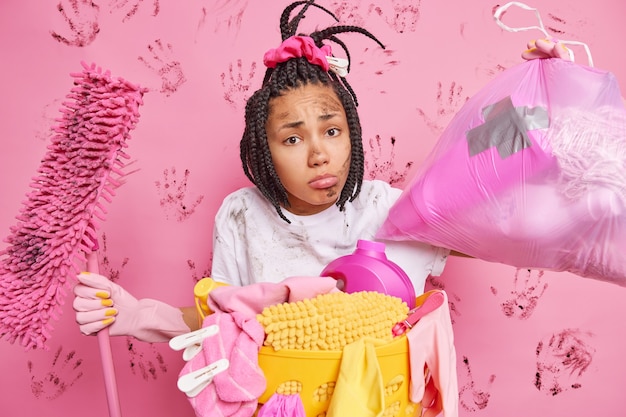 Mulher afro-americana descontente com dreadlocks carrega saco de lixo faz poses de esfregão perto do cesto de roupa suja dá dicas de limpeza ocupada fazendo trabalhos domésticos durante o fim de semana