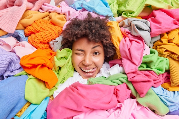 Mulher afro-americana divertida positiva cercada por roupas coloridas usadas coletadas para reciclagem ou doação e limpa seu guarda-roupa fica de frente para a pilha de roupas multicoloridas mostra a língua