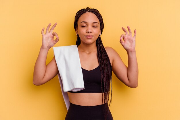 Mulher afro-americana do esporte jovem segurando uma toalha isolada na parede amarela relaxa após um árduo dia de trabalho, ela está realizando ioga.