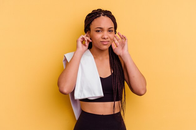 Mulher afro-americana do esporte jovem segurando uma toalha isolada no fundo amarelo, cobrindo as orelhas com as mãos.
