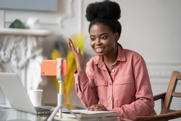 Mulher afro-americana do milênio com controle remoto de penteado afro, estudando, trabalhando online no laptop, conversando com amigos através de videochamada em smartphone no tripé. Blogger de gravação de blog de influenciador do Blogger.