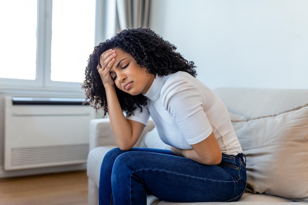 Mulher afro-americana em expressão dolorosa, segurando as mãos contra a barriga, sofrendo dor no período menstrual, deitada triste na cama em casa com cãibras na barriga no conceito de saúde feminina