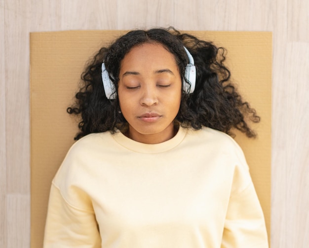 Mulher afro-americana em fones de ouvido descansando na esteira com os olhos fechados relaxando após o exercício