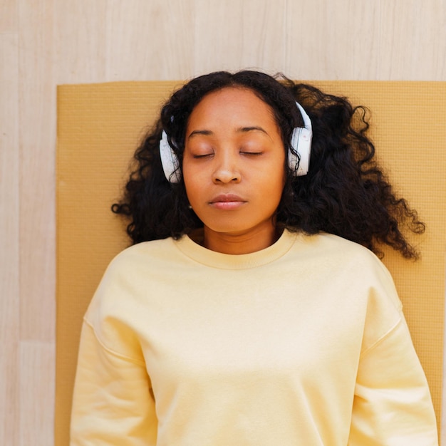 Mulher afro-americana em fones de ouvido descansando no tapete com os olhos fechados relaxando após o exercício