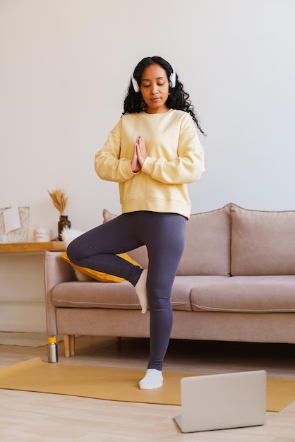 Mulher afro-americana em fones de ouvido em pé na árvore pose yoga enquanto segue o tutorial no laptop