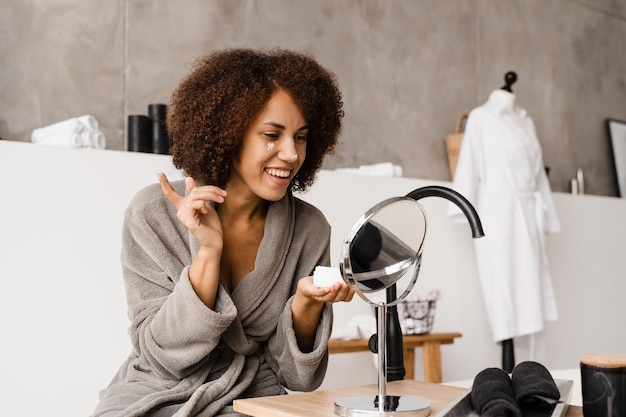 Mulher afro-americana em roupão de banho com creme hidratante facial fazendo rotina de beleza matinal Menina africana aplicando creme hidratante facial para proteger a pele da secura no banho