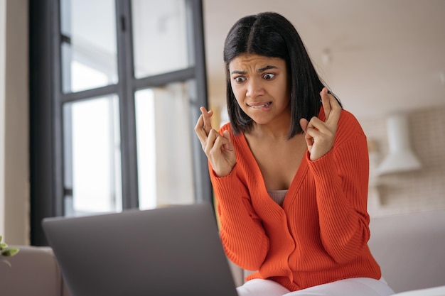 Mulher afro-americana emocional usando laptop segurando dedos cruzados fazendo compras on-line deseja algo