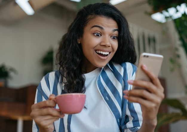 Mulher afro-americana empolgada usando um celular e assistindo a um vídeo em casa