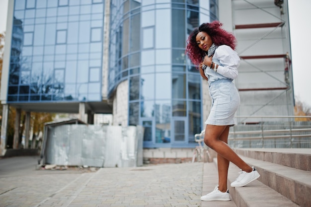 Mulher afro-americana encaracolada atraente em vestido jeans posou contra o edifício moderno de vários andares